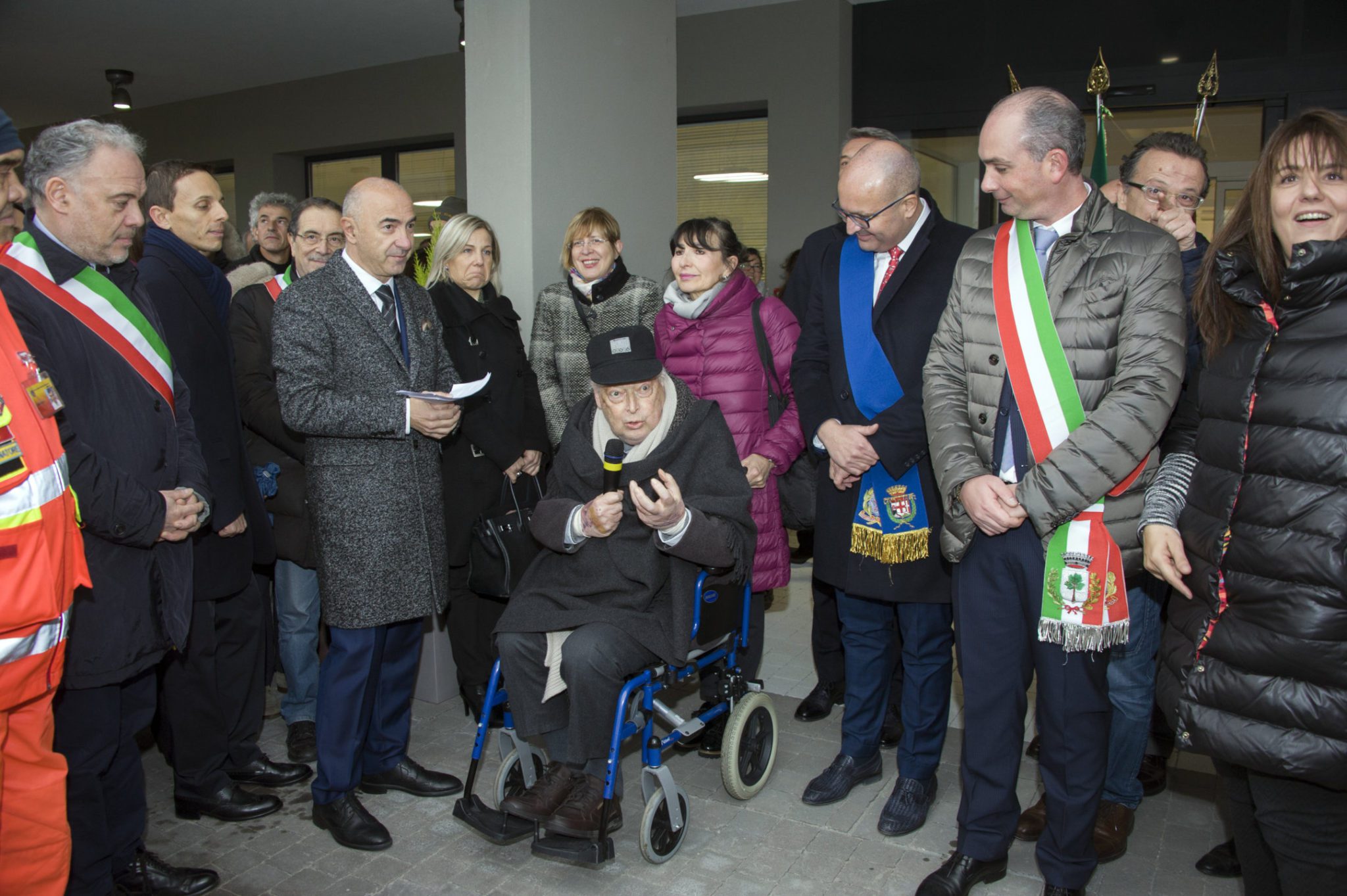 Un'immagine che ritrae sedia a rotelle, pantaloni, capispalla, gruppo sociale e cappotto.