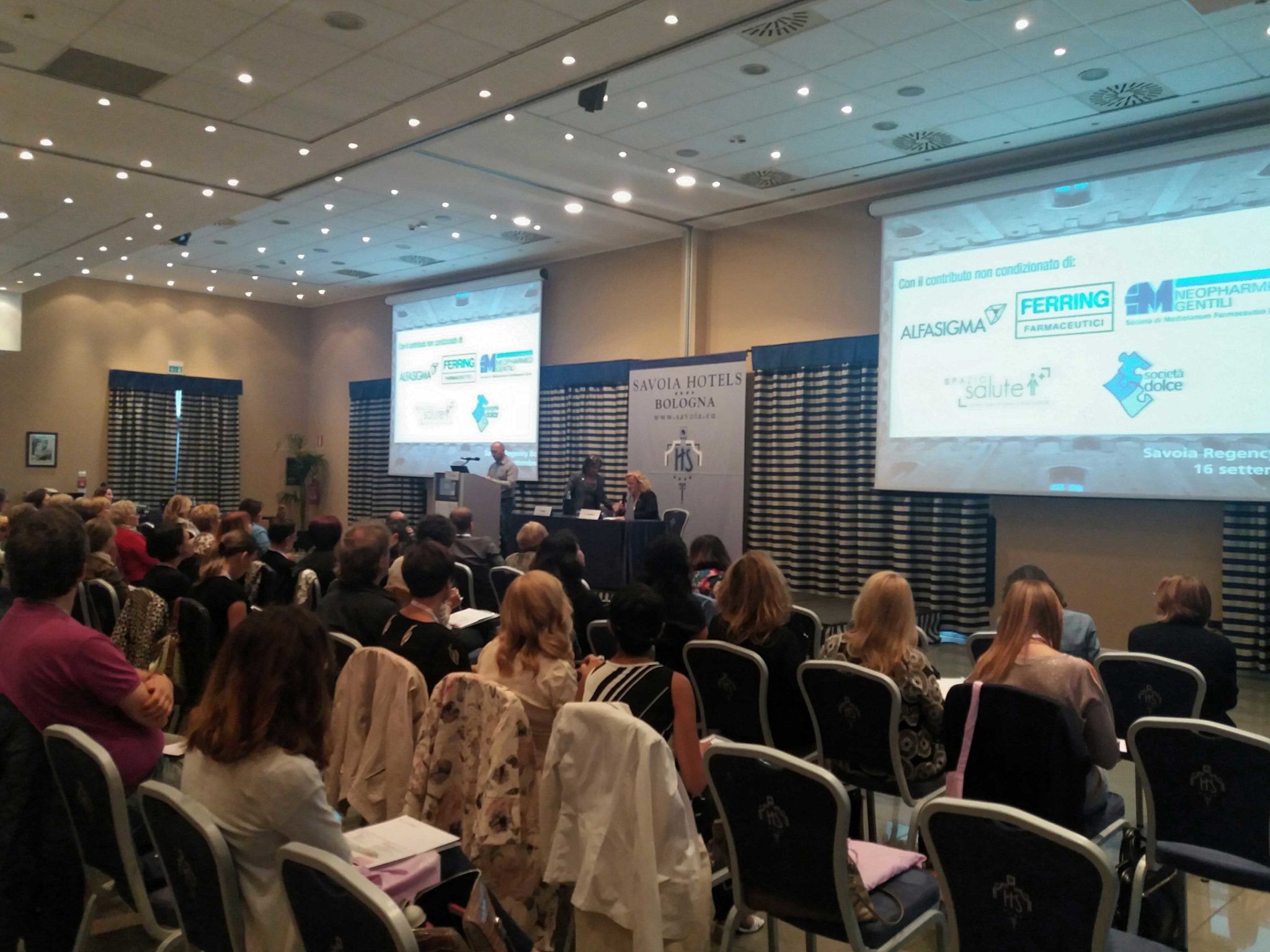 Un'immagine che ritrae mobilia, sedia, evento, dispositivo elettronico e dispositivo di visualizzazione.